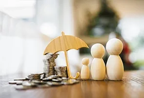 Three wooden figurines of varying sizes stand next to stacks of coins and a small umbrella, symbolizing financial protection and family savings