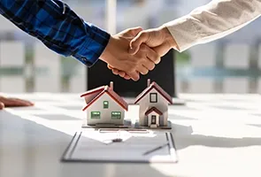 Two people are shaking hands over a table with small house models and a document, possibly finalizing a real estate deal